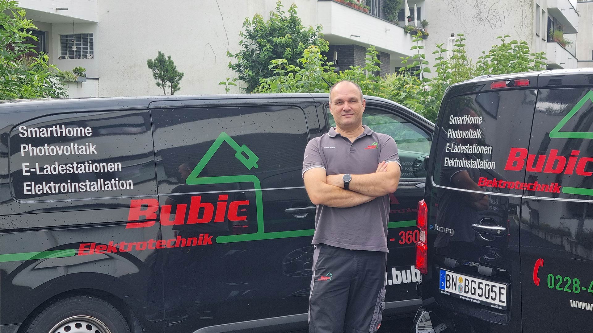 Ein Mann mit kurzem Haar und neutralem Gesichtsausdruck steht vor zwei schwarzen Firmenfahrzeugen