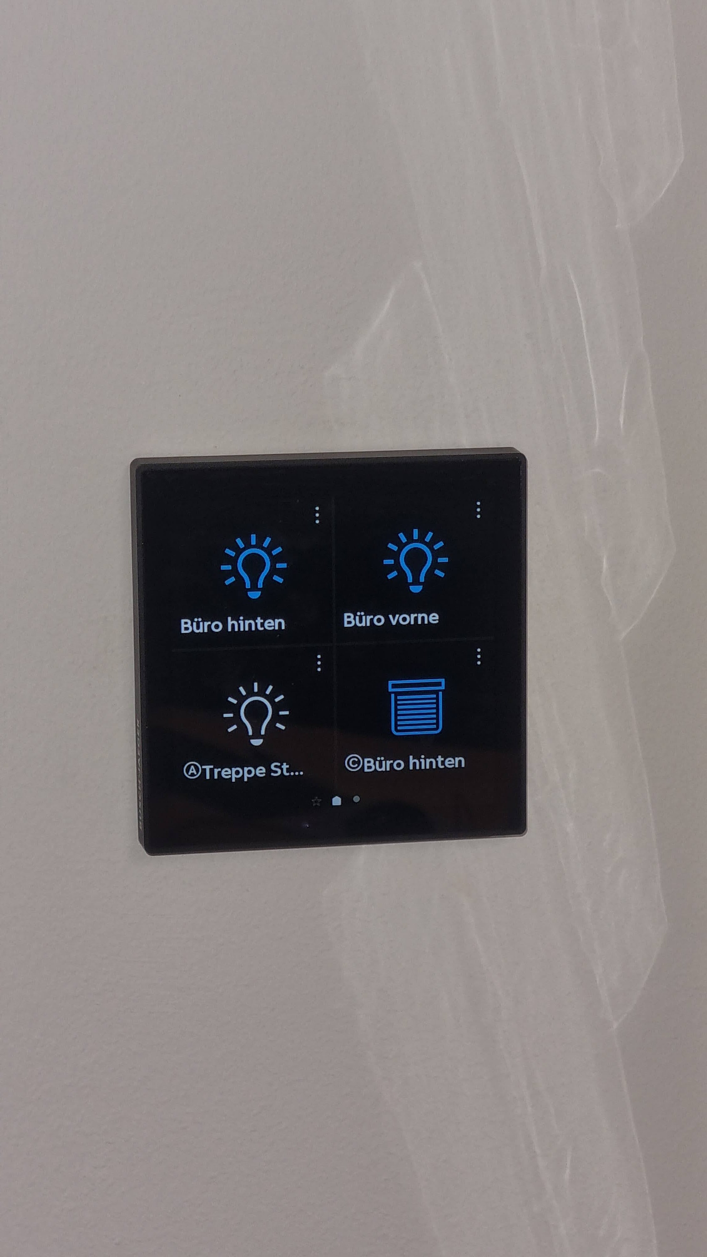 Ein schwarzes Bedienfeld mit blauen Lichtsymbolen und deutschen Beschriftungen an der Wand