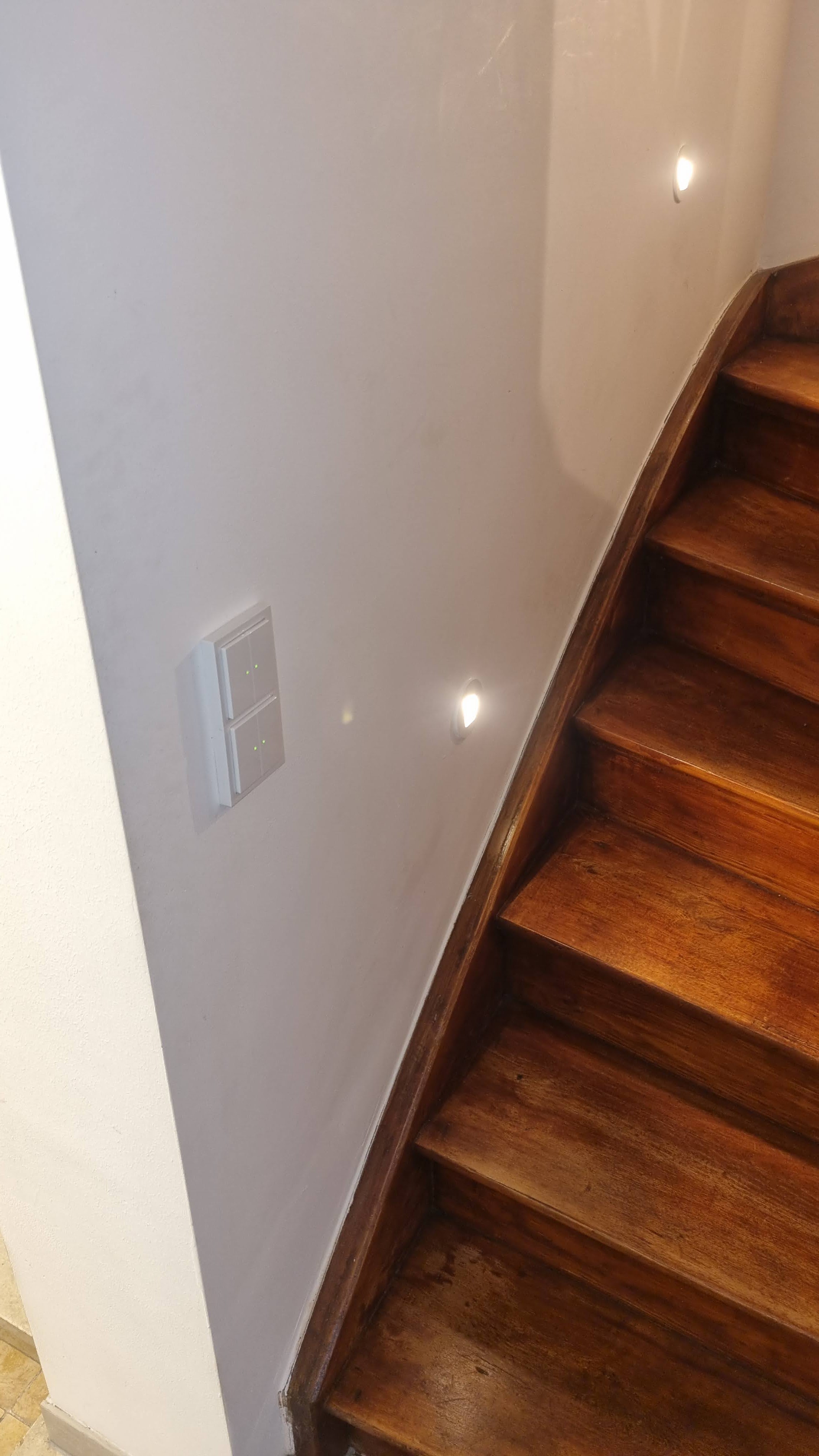 Eine Holztreppe mit Wandlichtern und einem weißen Wandschalter an heller Wand