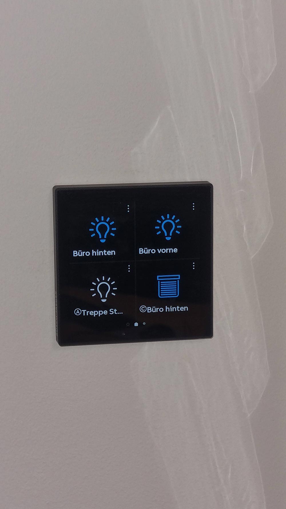 Schwarzes Touchpanel mit blauen Lichtsymbolen und Raumlabels an grauer Wand